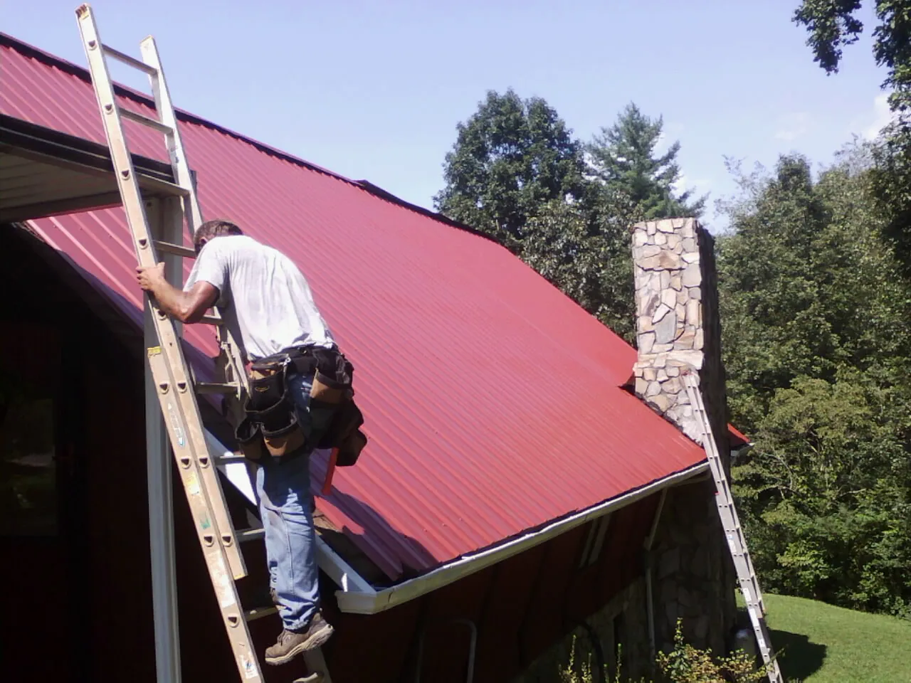 Roofer on a ladder beside a red metal roof and stone chimney — Soffit & Fascia Repair by Roofing Brothers in Ridgefield